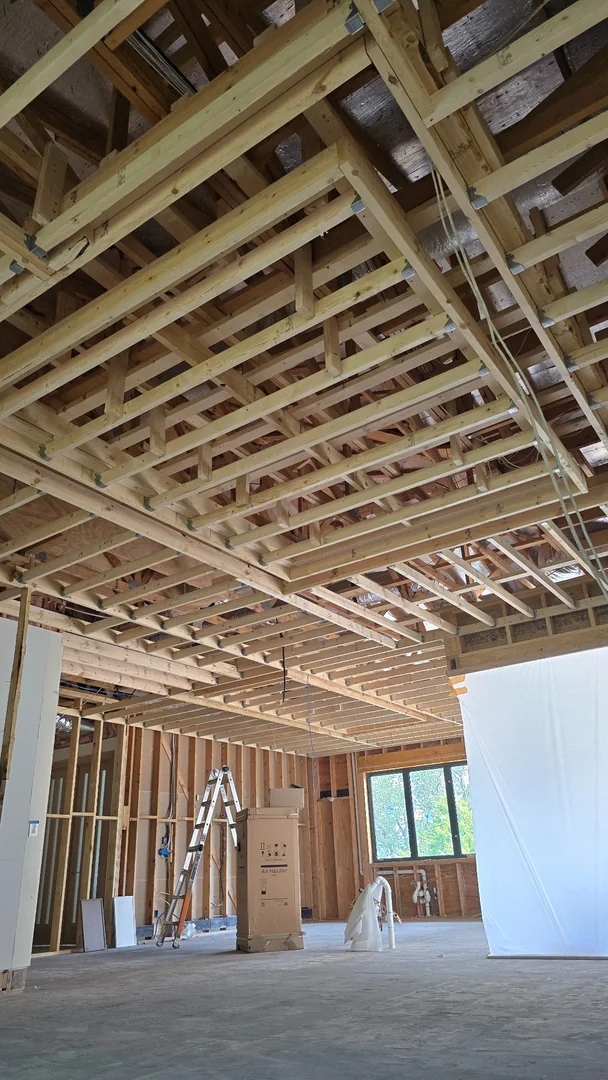 Exposed wood ceiling framing and blocking above an unfinished room, with electrical wiring, framed walls and a ladder.